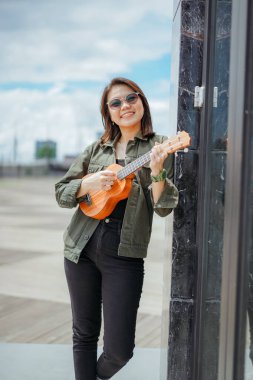 Ceket ve açık havada siyah kot giyen güzel bir Asyalı kadının Ukulele 'sini oynuyorum.