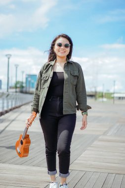Ceket ve açık havada siyah kot giyen güzel bir Asyalı kadının Ukulele 'sini oynuyorum.