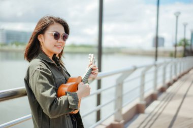 Ceket ve açık havada siyah kot giyen güzel bir Asyalı kadının Ukulele 'sini oynuyorum.