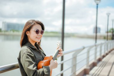 Ceket ve açık havada siyah kot giyen güzel bir Asyalı kadının Ukulele 'sini oynuyorum.