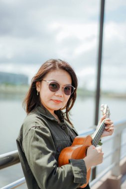 Ceket ve açık havada siyah kot giyen güzel bir Asyalı kadının Ukulele 'sini oynuyorum.