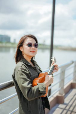 Ceket ve açık havada siyah kot giyen güzel bir Asyalı kadının Ukulele 'sini oynuyorum.