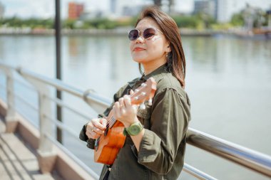 Ceket ve açık havada siyah kot giyen güzel bir Asyalı kadının Ukulele 'sini oynuyorum.