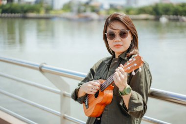 Ceket ve açık havada siyah kot giyen güzel bir Asyalı kadının Ukulele 'sini oynuyorum.