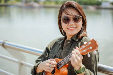 Ceket ve açık havada siyah kot giyen güzel bir Asyalı kadının Ukulele 'sini oynuyorum.