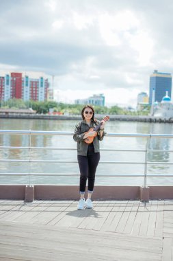 Ceket ve açık havada siyah kot giyen güzel bir Asyalı kadının Ukulele 'sini oynuyorum.