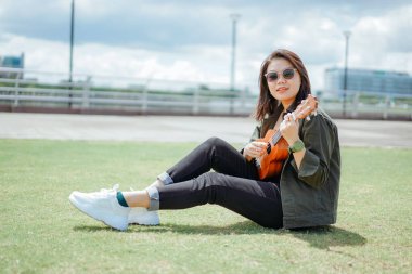 Ceket ve açık havada siyah kot giyen güzel bir Asyalı kadının Ukulele 'sini oynuyorum.