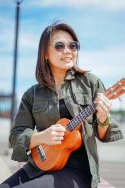 Ceket ve açık havada siyah kot giyen güzel bir Asyalı kadının Ukulele 'sini oynuyorum.