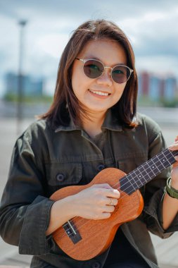 Ceket ve açık havada siyah kot giyen güzel bir Asyalı kadının Ukulele 'sini oynuyorum.