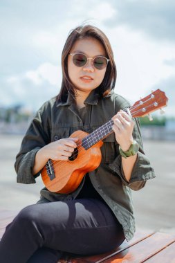 Ceket ve açık havada siyah kot giyen güzel bir Asyalı kadının Ukulele 'sini oynuyorum.