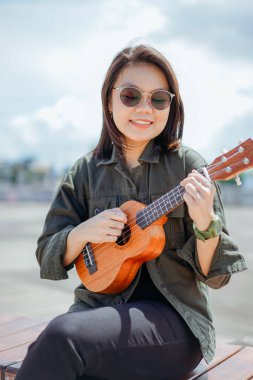 Ceket ve açık havada siyah kot giyen güzel bir Asyalı kadının Ukulele 'sini oynuyorum.