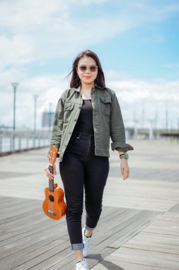 Ceket ve açık havada siyah kot giyen güzel bir Asyalı kadının Ukulele 'sini oynuyorum.
