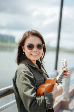Ceket ve açık havada siyah kot giyen güzel bir Asyalı kadının Ukulele 'sini oynuyorum.