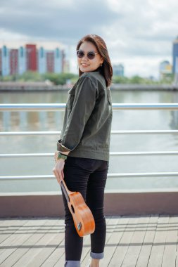 Ceket ve açık havada siyah kot giyen güzel bir Asyalı kadının Ukulele 'sini oynuyorum.