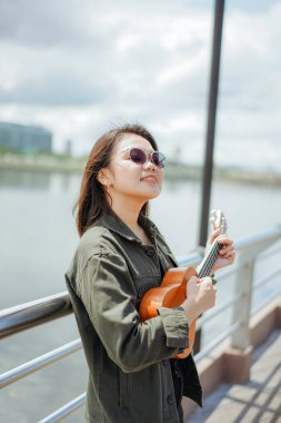 Ceket ve açık havada siyah kot giyen güzel bir Asyalı kadının Ukulele 'sini oynuyorum.