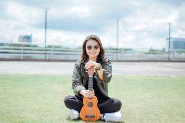 Ceket ve açık havada siyah kot giyen güzel bir Asyalı kadının Ukulele 'sini oynuyorum.
