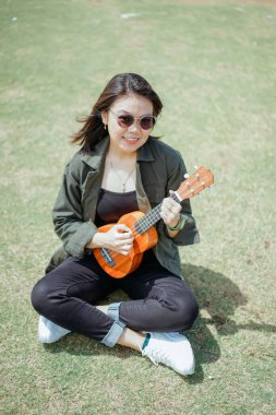 Ceket ve açık havada siyah kot giyen güzel bir Asyalı kadının Ukulele 'sini oynuyorum.