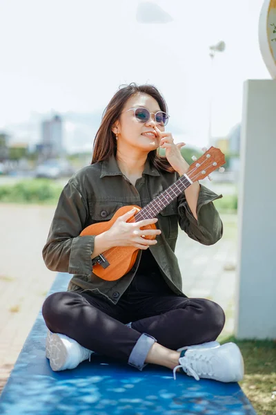 Ceket ve açık havada siyah kot giyen güzel bir Asyalı kadının Ukulele 'sini oynuyorum.