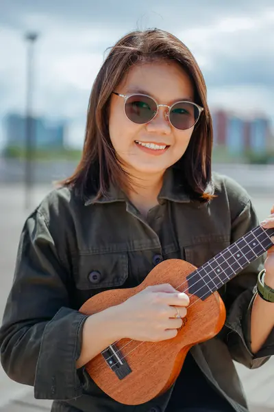Ceket ve açık havada siyah kot giyen güzel bir Asyalı kadının Ukulele 'sini oynuyorum.
