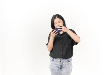 Genç Asyalı kadın, beyaz arka planda siyah tişört ve kot pantolon giyerek akıllı telefondan oyun oynuyor.