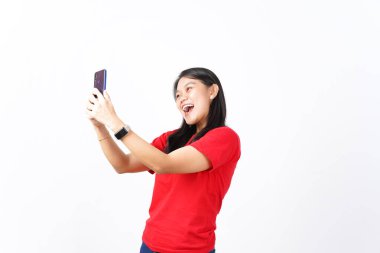 Güzel Asyalı kadın kamerayla selfie çekerken gülümsüyor, beyaz arka planda akıllı bir telefon tutuyor.