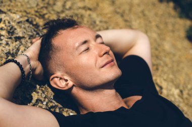 Genç adam gözlerini taşın üzerine kapatarak dinleniyor. Yakın plan portre.