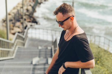 Düşünceli genç adam aşağı bakıyor, denize açılan merdivenlerde duruyor. Siyah tişört giymiş, güneş gözlüğü takıyor.