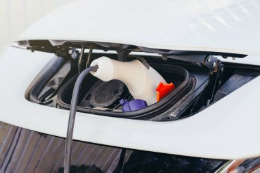 Beyaz elektrikli bir arabaya yakın çekim yaptırıyorum. Şarj portuna kablo takılıyor.