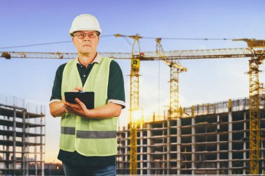 Foreman, 50 yaşlarında koruyucu bir miğfer ve işaret yeleği giyen bir adam tabletle bir inşaat alanının arka planına bakıyor..
