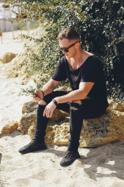 Siyah kotlu ve tişörtlü genç bir adam sahilde bir taşın üzerinde otururken telefona bir mesaj yazıyor..