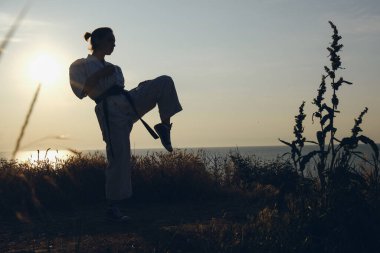 Bir kızın silueti okyanus üzerinde şafak güneşinin arka planına tekme atıyor. Karate, dövüş sanatları.