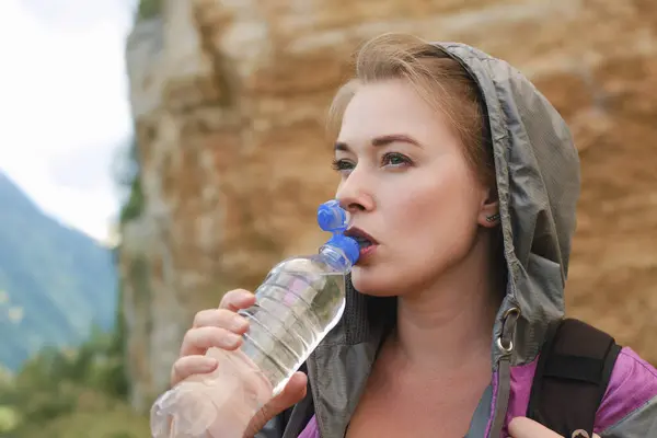 一个穿着风衣的旅游女孩在远足时喝瓶子里的水 — 图库照片