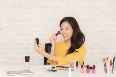 Gülümseyen Asyalı kadın makyaj masasında güzellik ürünleriyle otururken yüzünü kızartıyor..