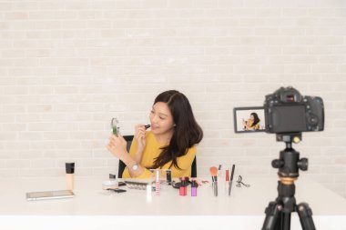 Gülümseyen Asyalı kadın, beyaz bir masanın üzerinde çeşitli makyaj ürünleriyle bir kameranın önüne ruj sürerek güzellik dersi kaydediyor..