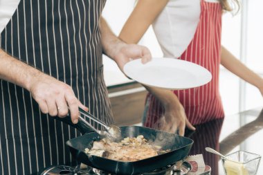 Yakın plan bir çift, ikisi de önlük giyiyorlar, tavada et pişiriyorlar. Biri tavadan yükselen buharlı bir tabak tutarken, diğeri maşa kullanıyor. Bu da sıcak ve lezzetli bir yemek öneriyor..