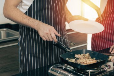 Yakın plan bir çift, ikisi de önlük giyiyorlar, tavada et pişiriyorlar. Biri tavadan yükselen buharlı bir tabak tutarken, diğeri maşa kullanıyor. Bu da sıcak ve lezzetli bir yemek öneriyor..