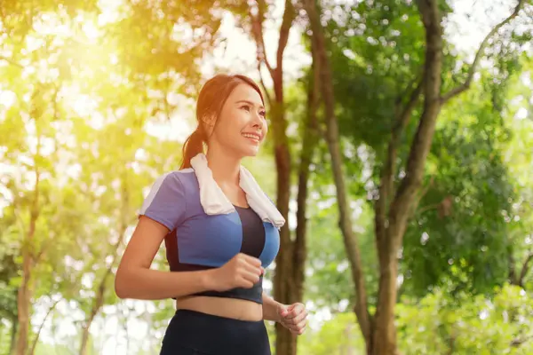 Boynunda havluyla gülümseyen Asyalı kadın parkta koşuyor, neşe ve aktif yaşam tarzını somutlaştırıyor..