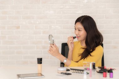 Gülümseyen Asyalı kadın, beyaz bir masanın üzerinde çeşitli makyaj ürünleriyle bir kameranın önüne ruj sürerek güzellik dersi kaydediyor..