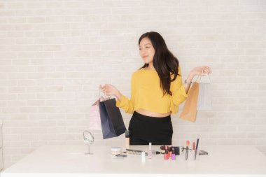 Masasında makyaj malzemeleri olan heyecanlı bir Asyalı kadın başarılı bir güzellik nakliyatından sonra neşe içinde alışveriş torbalarını tutuyor..