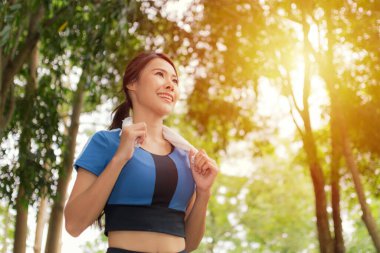 Boynunda havluyla gülümseyen Asyalı kadın parkta koşuyor, neşe ve aktif yaşam tarzını somutlaştırıyor..