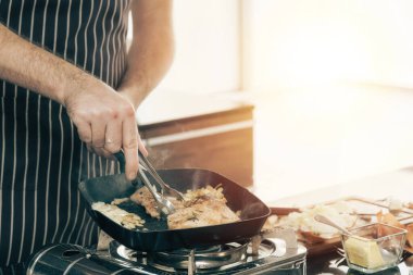 Yakın plan bir çift, ikisi de önlük giyiyorlar, tavada et pişiriyorlar. Biri tavadan yükselen buharlı bir tabak tutarken, diğeri maşa kullanıyor. Bu da sıcak ve lezzetli bir yemek öneriyor..
