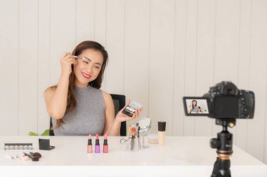 Neşeli genç Asyalı kadın kameraya gülümserken kaşlarını tımar etmek için fırça kullanıyor, elinde küçük bir ayna tutuyor..