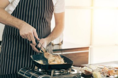 Yakın plan bir çift, ikisi de önlük giyiyorlar, tavada et pişiriyorlar. Biri tavadan yükselen buharlı bir tabak tutarken, diğeri maşa kullanıyor. Bu da sıcak ve lezzetli bir yemek öneriyor..