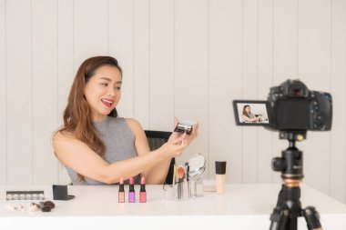 Gülümseyen Asyalı güzellik etkileyicisi, masasında çeşitli kozmetik ürünleri olan bir makyaj ürününü kameraya sunan bir video kaydeder..