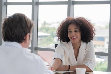 Çeşitli iş dallarında uzman bir erkek ve Afrikalı bir kadın, anlaşma ya da ortaklığı simgeleyen modern parlak bir ofiste el sıkışıyorlar..