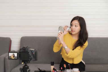 Genç Asyalı bir kadın kamerayla bir güzellik videosu kaydediyor. Kanepede aynaya bakarken kirpiklerini nasıl kıvıracağını gösteriyor..