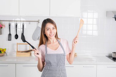 Güzel Asyalı genç kadın beyaz mutfağında neşe içinde poz veriyor, elinde yemek pişirme aleti ev yapımı yemeğe olan aşkı temsil eden bir spatula ve tahta kaşık..