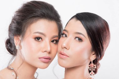 Yumuşak makyajlı ve karmaşık mücevherleri olan iki güzel Asyalı kadın, beyaz arka planda stüdyo çekimlerinde zarafeti sembolize ediyorlar..