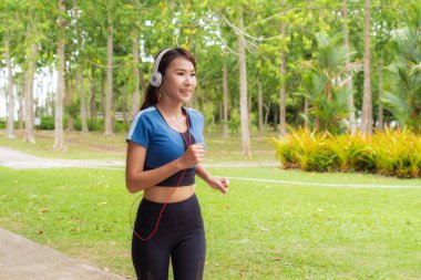 Gülümseyen genç Asyalı kadın yemyeşil parkta kulaklıkla müzik dinleyerek ferahlatıcı koşudan keyif alıyor. Sağlık ve refah için mükemmel.