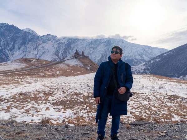 Asian tourist man in winter clothes standing in front of landscape view of Gergeti Holy Trinity Church on hill and Caucasus mountains - Tsminda Sameba. Beautiful Georgian landmark in early winter.